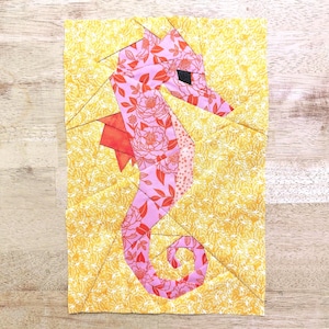 May include: A fabric art piece featuring a seahorse design. The seahorse is pink with a floral pattern and orange accents, set against a yellow background with a subtle pattern. The artwork is displayed on a wooden surface.