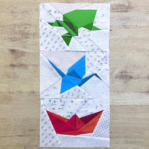 May include: Three fabric blocks with origami designs: a green frog, a blue crane, and a red and orange boat. The blocks are white with a small pattern and are arranged vertically.