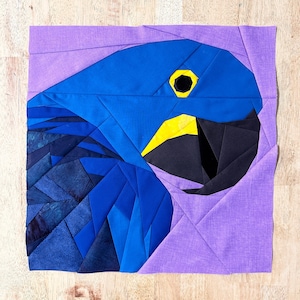 May include: A blue and black fabric patchwork design of a parrot's head with a yellow beak and a black eye. The parrot's head is on a purple background.