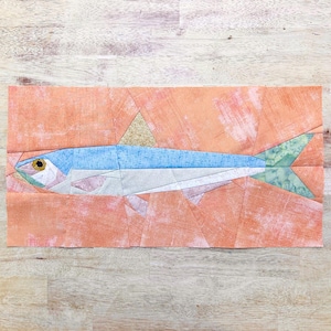 Puede incluir: Una pieza de arte textil que presenta un diseño de pez estilizado. El pez tiene un cuerpo azul, vientre blanco y una aleta caudal verde estampada. Se encuentra sobre un fondo naranja texturizado con una aleta triangular dorada. La obra de arte es probablemente un bloque de edredón o arte textil.