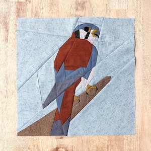 May include: A fabric art piece featuring a geometric design of a bird of prey perched on a branch. The bird has a brown body, blue wings, and a white chest. The background is light blue, and the branch is brown. The artwork is square.
