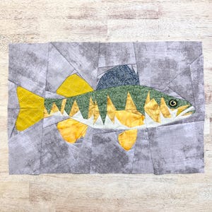 May include: A patchwork quilt depicting a fish with yellow, green, and white scales. The fish is set against a gray background.