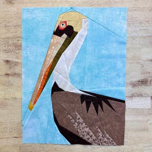 May include: A brown and white paper pieced pelican with a long orange beak and a blue background. The pelican is looking to the right.