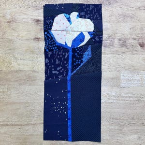 May include: A fabric flower applique with a white and blue flower design. The flower is made of various patterned fabrics, including polka dots, hearts, and floral prints. The stem is a solid blue with a blue stripe down the center. The background is a light wood grain.