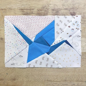 May include: A rectangular fabric block with a geometric design. The central shape is a vibrant blue, surrounded by white fabric with small patterns of dots, bicycles, and paper airplanes. The block is likely part of a larger quilt or textile project.