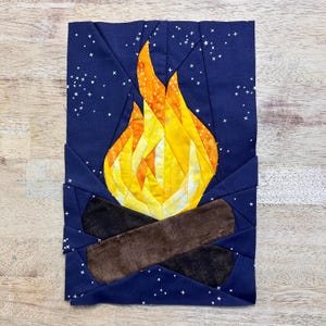 May include: A paper pieced quilt block depicting a campfire with a yellow and orange flame against a dark blue background with white stars.