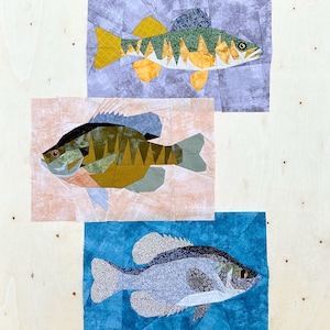 May include: Three different fish are shown in a patchwork style. The fish are made of fabric and are sewn together in a quilt-like pattern. The fish are a yellow and green bass, a brown and green sunfish, and a blue and black crappie.