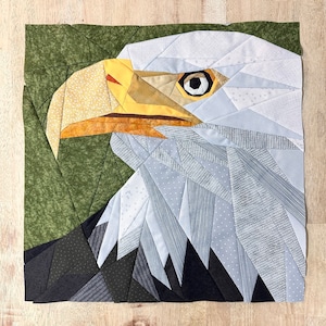 Könnte beinhalten: Eine weiße und graue Patchwork-Decke, die einen Weißkopfseeadler mit gelbem Schnabel und braunen Augen darstellt. Der Adler blickt nach links und hat einen grünen Hintergrund.