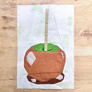 May include: A fabric quilt block featuring a pieced design of a caramel apple. The apple is rendered in shades of brown with a bright green top and a tan stick, set against a white background with subtle light green mottled texture throughout.