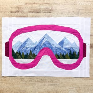 May include: A pink fabric patchwork design of a pair of ski goggles with a mountain scene inside. The goggles are made of pink fabric with a white fabric background. The mountain scene is made of blue, white, and green fabric.