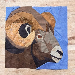 May include: A close-up of a quilt depicting a bighorn sheep's head. The quilt is made of various shades of brown, tan, and gray fabric. The sheep's eye is a bright orange color. The background is a light blue fabric.