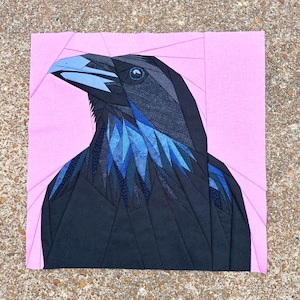 May include: A textile artwork depicting a raven's head and upper body. The raven is mainly black with blue detailing on the beak and chest. The background is a solid pink, and the piece is made from geometric fabric shapes.