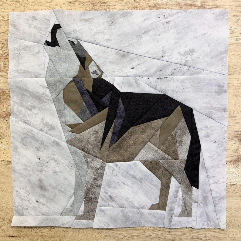 May include: A paper pieced quilt block featuring a wolf howling at the moon. The wolf is made of various shades of brown, black, and gray fabric. The background is a white fabric with a subtle gray marbling pattern.