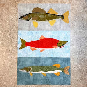 Può includere: Un blocco di trapunta con tre diversi disegni di pesci. Il pesce in alto è un pesce persico verde e marrone, il pesce al centro è un salmone rosso e il pesce in basso è un luccio verde e bianco. Lo sfondo è un tessuto blu e bianco con un motivo ondulato.