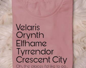 May include: A dusky pink t-shirt featuring the text "Velaris, Orynth, Elfhame, Tyrrendor, Crescent City" in black. The phrase "Oh, the places I'd like to go..." is printed below the list. The shirt is placed on a white, fluffy surface.