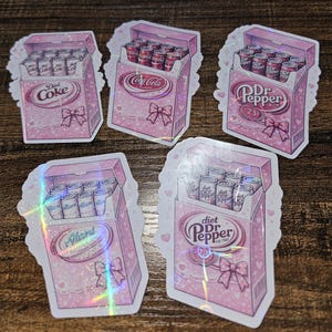 May include: A collection of pink stickers featuring soda can boxes. Each box has a bow and contains cans of Diet Coke, Coca-Cola, Dr. Pepper, and Alan's. The stickers have a holographic effect and are decorated with hearts and lace.