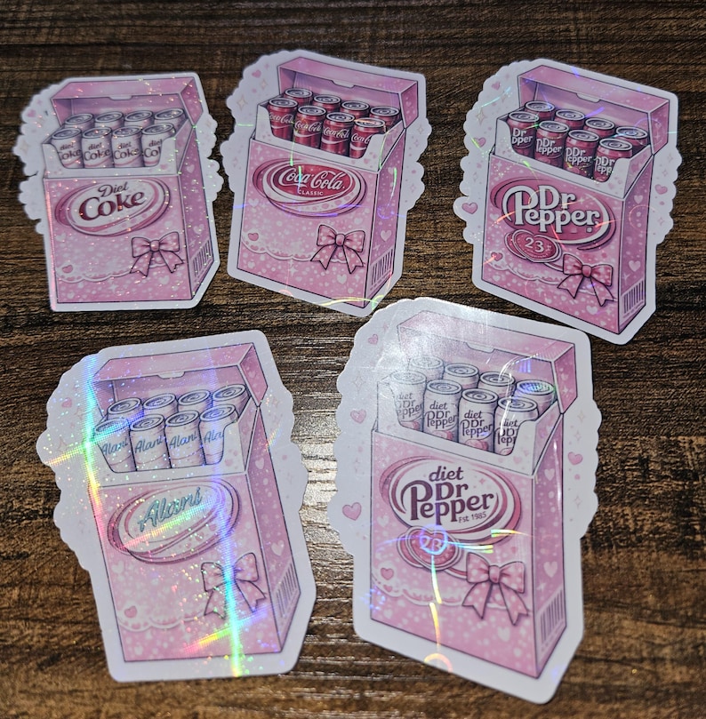 May include: A collection of pink stickers featuring soda can boxes. Each box has a bow and contains cans of Diet Coke, Coca-Cola, Dr. Pepper, and Alan's. The stickers have a holographic effect and are decorated with hearts and lace.
