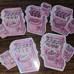 May include: Pink stickers featuring boxes of soda cans designed to look like cigarette packs. The boxes are decorated with bows and the soda brands Diet Coke, Coca-Cola, Dr. Pepper, and Alan's are visible.