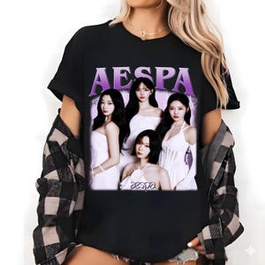 Może przedstawiać: Czarny t-shirt z fioletowo-białą grafiką przedstawiającą cztery kobiety w białych sukienkach, z napisem "AESPA" nad nimi. Słowo "aespa" jest również wydrukowane na dole obrazu.