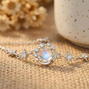 May include: A silver bracelet featuring a moon and star design. The bracelet has a central moon-shaped pendant with a round, iridescent stone surrounded by small, clear crystals. Star-shaped charms with crystals are spaced along the chain.