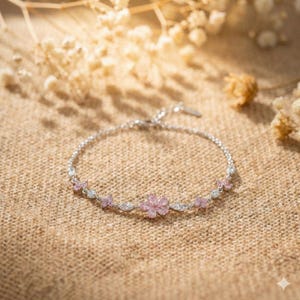 以下が含まれることがあります： ピンクとクリアな宝石をあしらった、繊細なシルバーブレスレット。ブレスレットの中央には花があり、その周りには小さなピンクの花とクリアなアクセントがあります。質感のあるニュートラルな色の表面に表示されています。