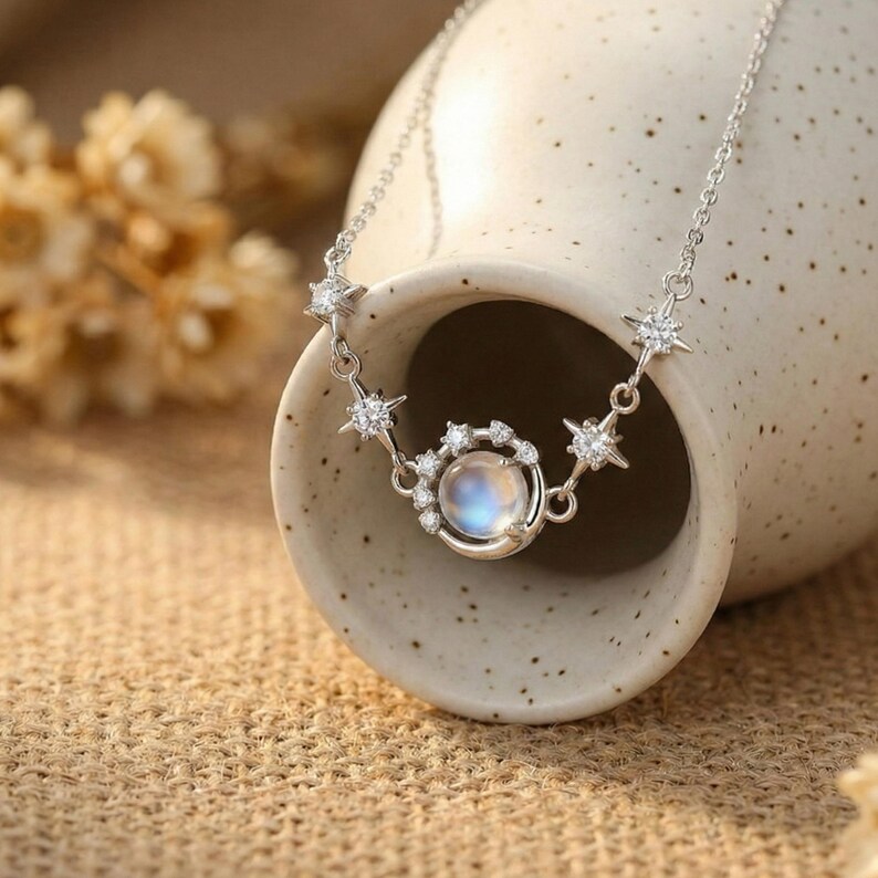 May include: A silver necklace featuring a celestial design. The necklace has a central pendant with a round, iridescent stone surrounded by smaller clear stones. Star-shaped accents with clear stones are spaced along the chain.