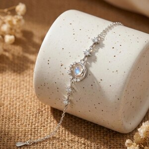 May include: A silver bracelet featuring a celestial design. The bracelet has a chain with star-shaped accents and a central pendant with a moonstone and a halo of small crystals. The bracelet is displayed on a neutral-colored surface.