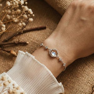 May include: A silver bracelet featuring a central moonstone pendant encircled by a heart-shaped frame. The bracelet is adorned with small star-shaped accents along the chain. The jewelry is displayed on a wrist, with a white ruffled sleeve in the background.