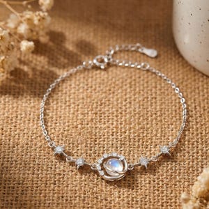 Könnte beinhalten: Ein silbernes Armband mit einem himmlischen Design. Das Armband hat eine feine Kette mit einem zentralen Anhänger, der einem Planeten ähnelt, umgeben von kleinen, funkelnden Kristallen. Sternförmige Akzente sind entlang der Kette angebracht.