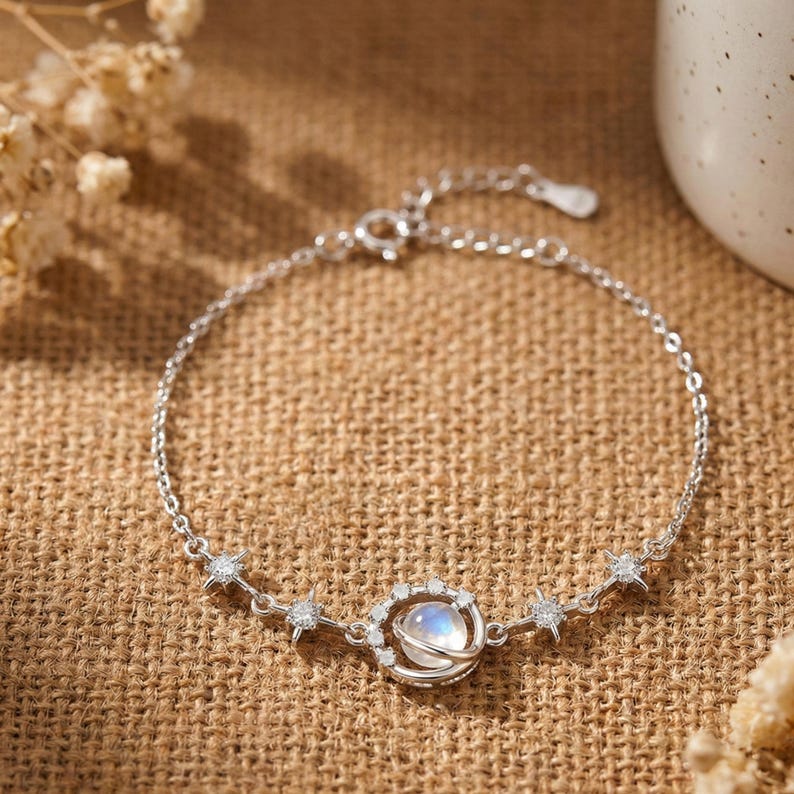 May include: A silver bracelet featuring a celestial design. The bracelet has a delicate chain with a central pendant resembling a planet, encircled by small, sparkling crystals. Star-shaped accents are placed along the chain.