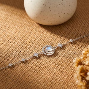 May include: A silver bracelet featuring a central moonstone orb encircled by a delicate metal frame. The bracelet is adorned with star-shaped and round clear gemstones, set against a textured, tan background.