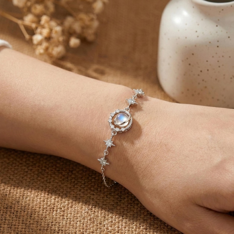 May include: A silver bracelet featuring a central moonstone surrounded by a halo of smaller stones. The bracelet is adorned with star-shaped accents and is worn on a wrist. The design is elegant and celestial.