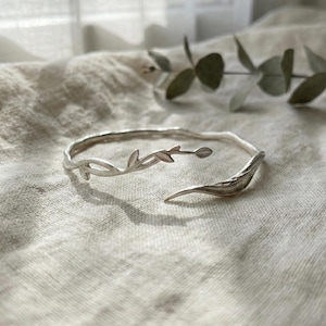 May include: A silver cuff bracelet with a botanical design. The open bracelet features a vine with leaves and a curved, leaf-shaped end. The bracelet is displayed on a textured, off-white fabric. The bracelet's design is reminiscent of a natural twig.