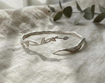 May include: A silver-colored cuff bracelet with a nature-inspired design. The open-ended bracelet features a delicate vine motif with leaves and a curved, leaf-shaped end. The bracelet is displayed on a textured, off-white fabric.