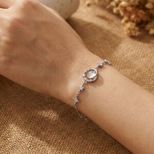 May include: A silver bracelet featuring a moonstone pendant surrounded by a halo of small, clear crystals. The bracelet also includes a series of star-shaped charms. The jewelry is displayed on a wrist against a neutral background.