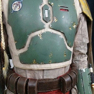 May include: A detailed costume of a bounty hunter, featuring a weathered green chest plate with a circular emblem. The costume includes a brown leather belt, red rope, and a tan undersuit. The armor has a distressed appearance.