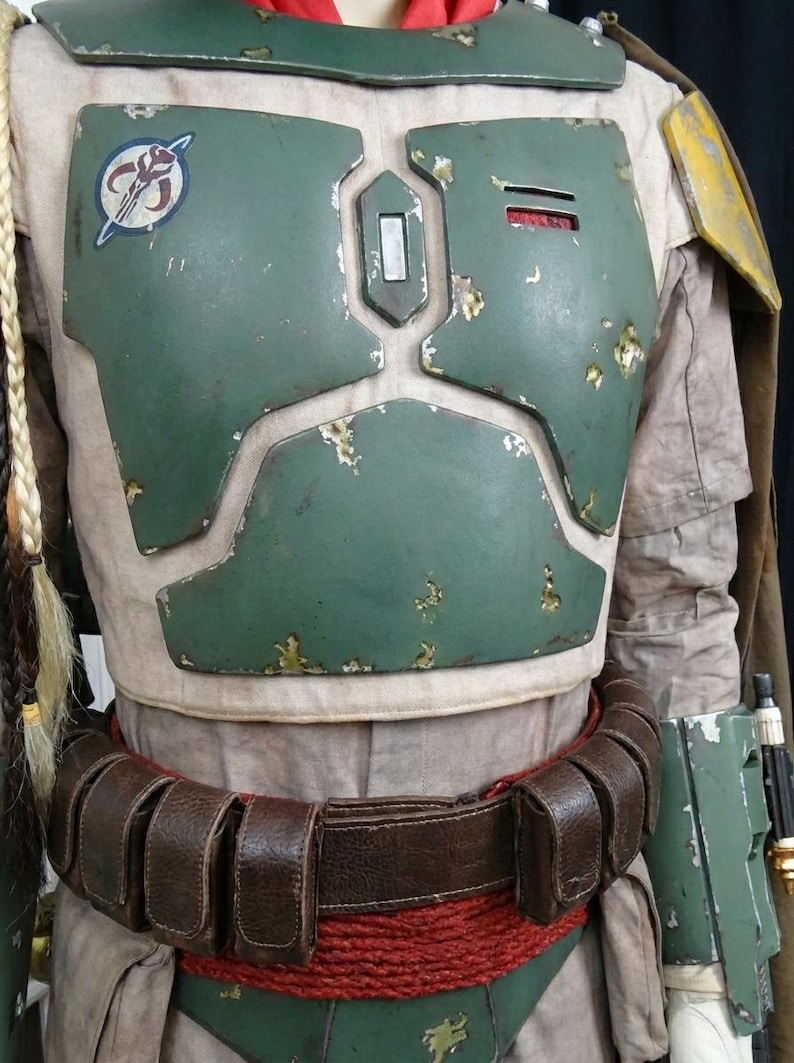 May include: A detailed costume of a bounty hunter, featuring a weathered green chest plate with a circular emblem. The costume includes a brown leather belt, red rope, and a tan undersuit. The armor has a distressed appearance.
