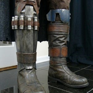 May include: Close-up of brown leather boots and armored leg coverings. The boots have a worn appearance with straps and buckles. The leg armor features metal plating and cylindrical details, suggesting a futuristic or fantasy theme.