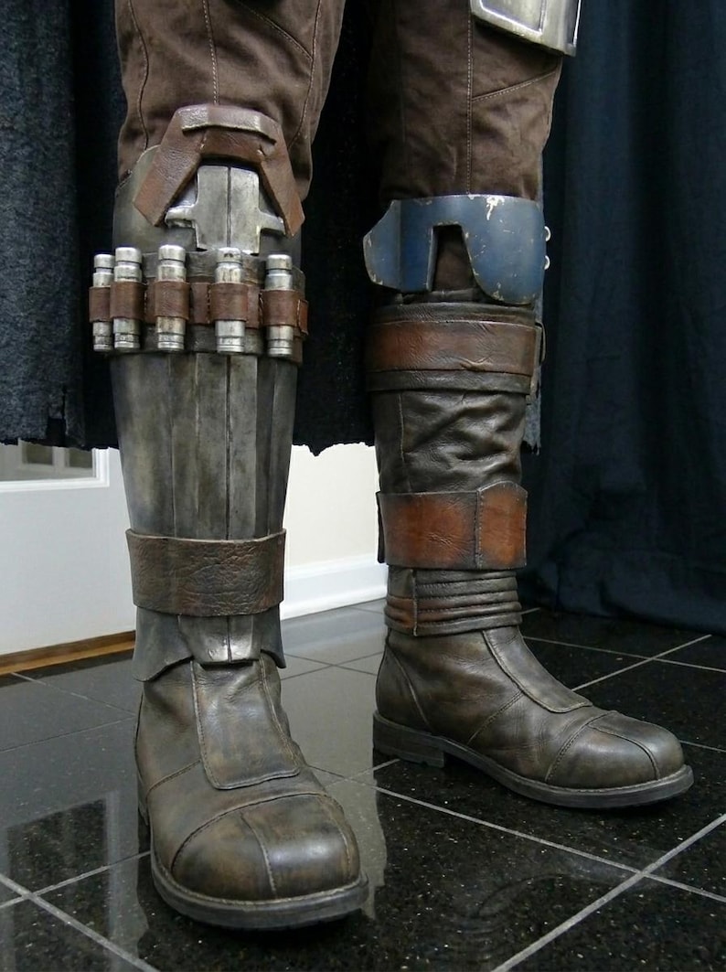 May include: Close-up of brown leather boots and armored leg coverings. The boots have a worn appearance with straps and buckles. The leg armor features metal plating and cylindrical details, suggesting a futuristic or fantasy theme.