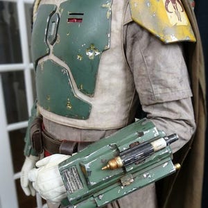 May include: A detailed costume featuring a green and yellow chest plate, shoulder armor with a symbol, and a green gauntlet with attached devices. The costume has a weathered appearance, suggesting use. A brown cape is visible.