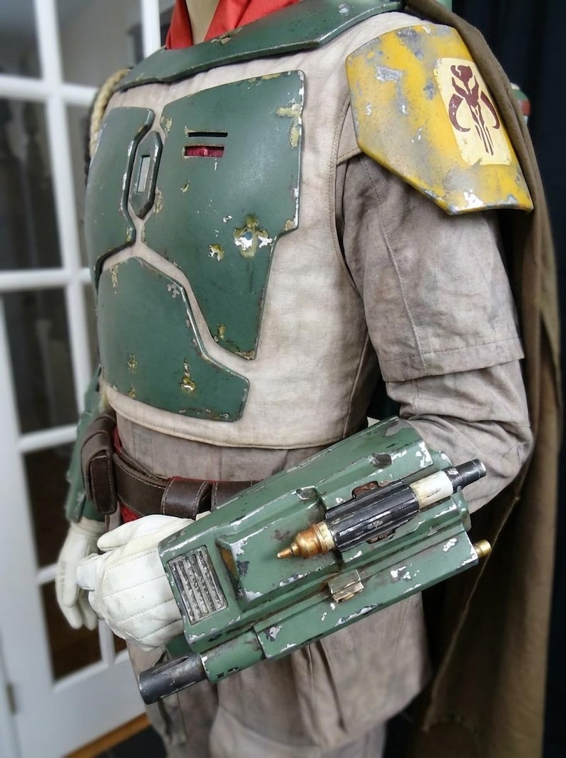 May include: A detailed costume featuring a green and yellow chest plate, shoulder armor with a symbol, and a green gauntlet with attached devices. The costume has a weathered appearance, suggesting use. A brown cape is visible.