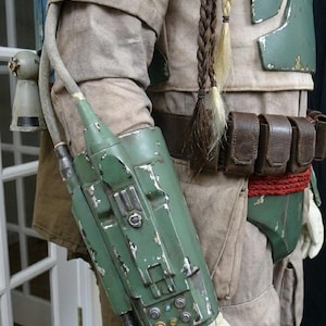 May include: A detailed costume element featuring a weathered green arm gauntlet with various buttons and a hose attachment. The costume includes a tan jacket, green armor, a brown belt with pouches, and braided hair extensions.
