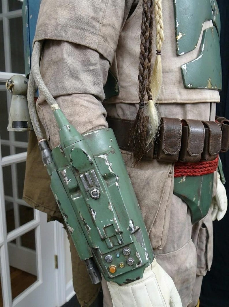 May include: A detailed costume element featuring a weathered green arm gauntlet with various buttons and a hose attachment. The costume includes a tan jacket, green armor, a brown belt with pouches, and braided hair extensions.
