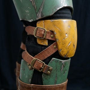 K&ouml;nnte beinhalten: Nahaufnahme eines gr&uuml;n-gelben, verwitterten R&uuml;stungsteils mit braunen Lederriemen und Schnallen. Die R&uuml;stung scheint Teil eines Kost&uuml;ms zu sein, mit sichtbaren Gebrauchsspuren, was auf eine kampferprobte &Auml;sthetik hindeutet.