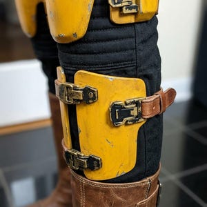 K&ouml;nnte beinhalten: Nahaufnahme von gelb-schwarzen R&uuml;stungsteilen mit Metallschnallen und braunen Lederriemen. Die R&uuml;stung wird &uuml;ber einer schwarzen Hose und braunen Lederstiefeln getragen. Die R&uuml;stung hat ein abgenutztes Aussehen.