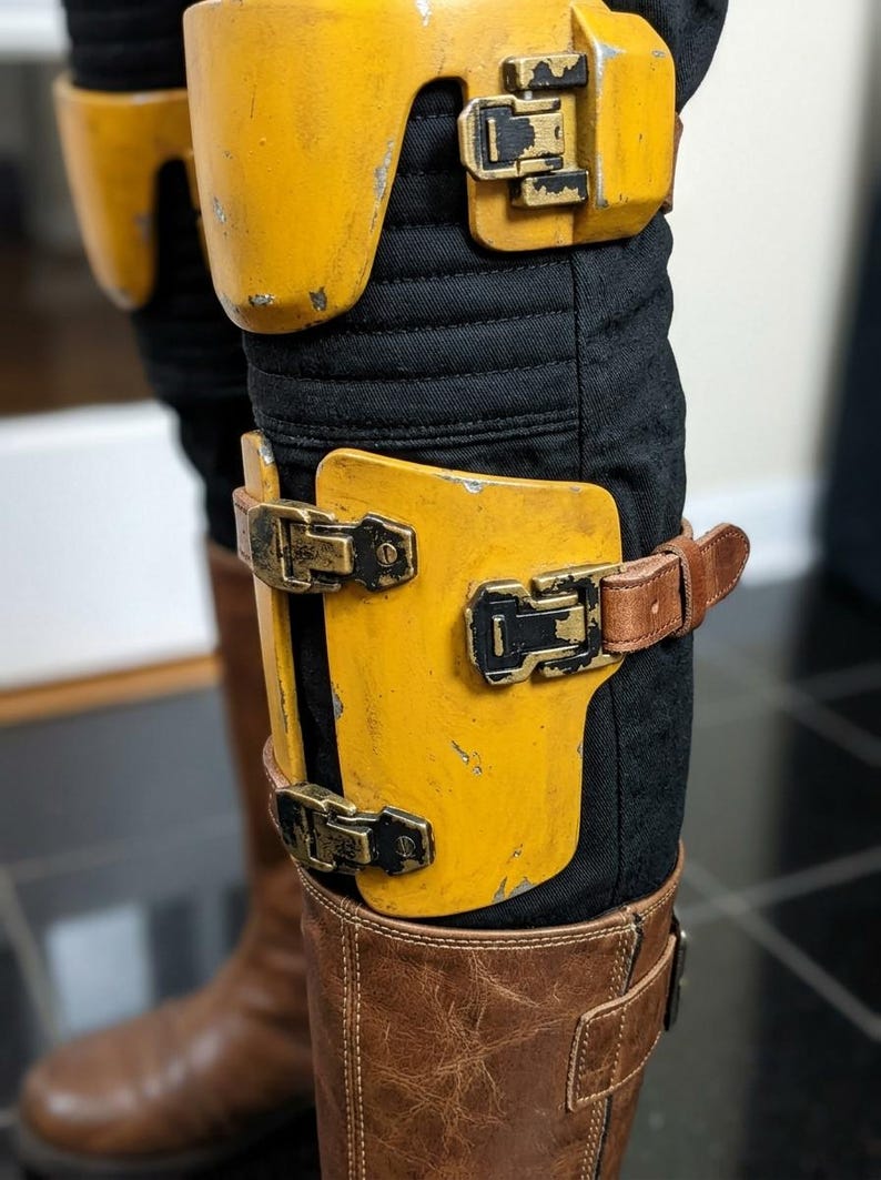K&ouml;nnte beinhalten: Nahaufnahme von gelb-schwarzen R&uuml;stungsteilen mit Metallschnallen und braunen Lederriemen. Die R&uuml;stung wird &uuml;ber einer schwarzen Hose und braunen Lederstiefeln getragen. Die R&uuml;stung hat ein abgenutztes Aussehen.