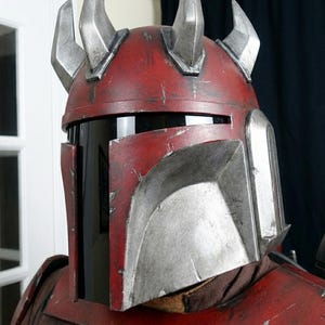 Puede incluir: Un casco rojo y plateado desgastado con cuatro cuernos plateados. El casco tiene una visera negra y forma parte de un disfraz. El casco tiene una apariencia desgastada con ara&ntilde;azos y desgaste.