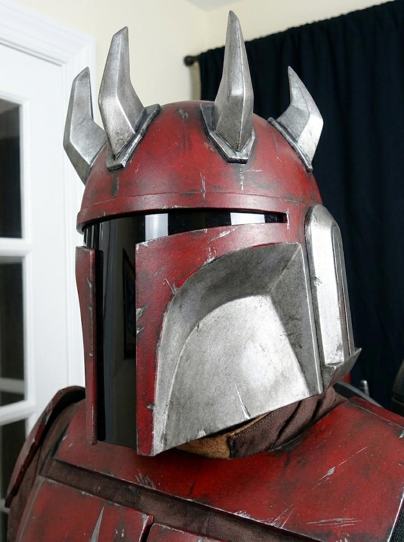 Puede incluir: Un casco rojo y plateado desgastado con cuatro cuernos plateados. El casco tiene una visera negra y forma parte de un disfraz. El casco tiene una apariencia desgastada con ara&ntilde;azos y desgaste.