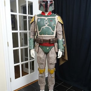 May include: A full-body costume resembling the character Boba Fett from Star Wars. The costume features a green and brown helmet, a green and brown chest plate, and tan pants. The costume includes a cape, belt, and white gloves and boots.