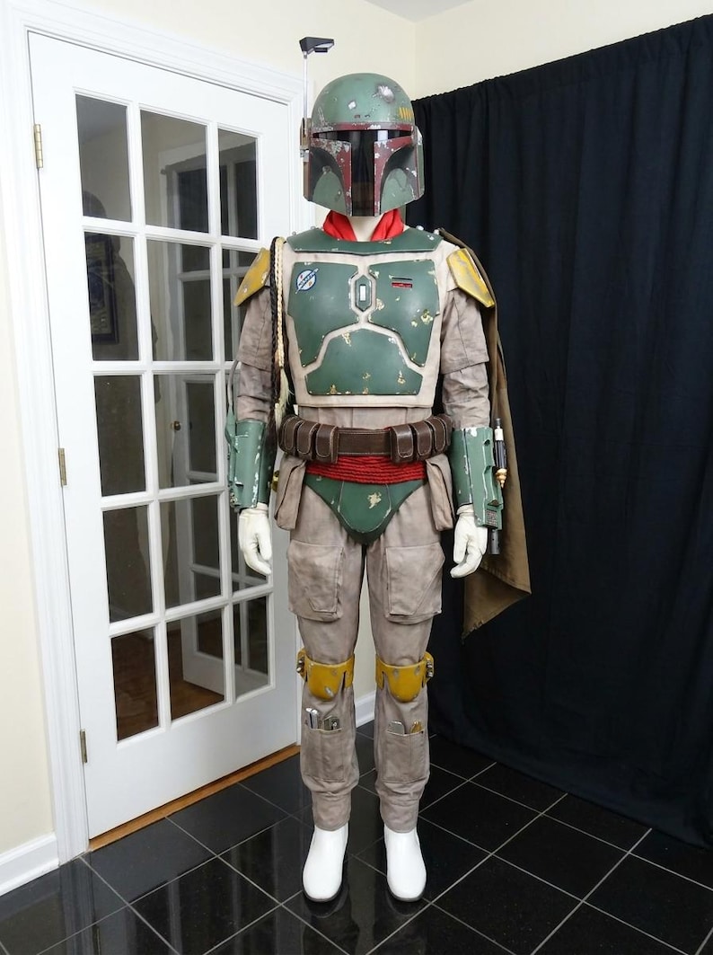 May include: A full-body costume resembling the character Boba Fett from Star Wars. The costume features a green and brown helmet, a green and brown chest plate, and tan pants. The costume includes a cape, belt, and white gloves and boots.